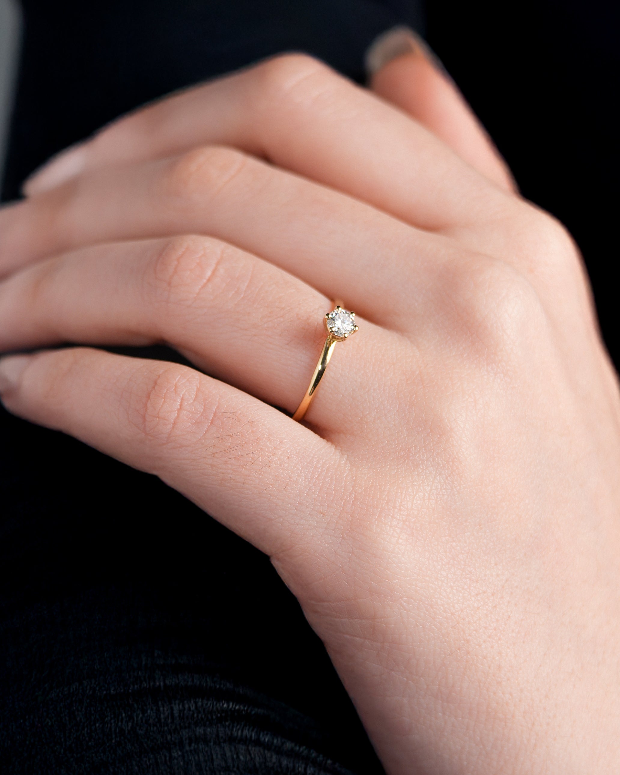 Anel de Noivado Amélie I Solitário em Ouro 18K Amarelo com Moissanite de 0,25ct (4mm) em uma mão branca em cima de uma blusa preta.